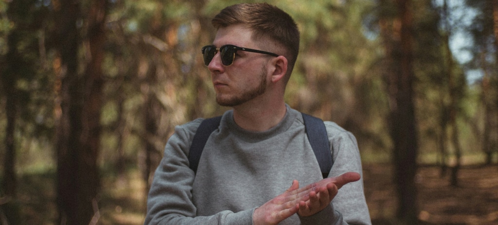 A young man with short blonde hair walks through a forest, rubbing his hands together. He wears black Ray-Ban Clubmaster sunglasses, a grey sweater, and a backpack on his back.