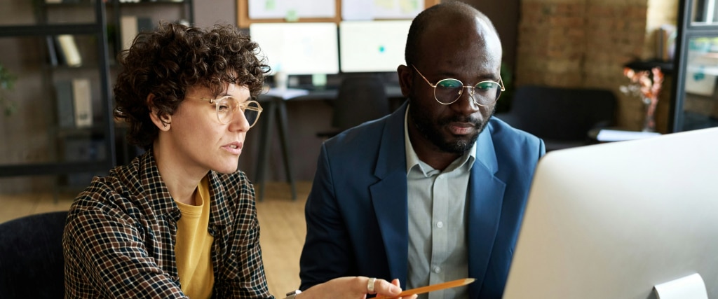 In a bright, modern office, two colleagues collaborate in front of a Mac computer. The woman, with short brown curly hair, wearing transparent beige round glasses and a mustard t-shirt layered under a check shirt, gestures toward the screen with a pencil. Next to her, a bald man with a tidy beard, dressed in a navy suit and light blue shirt, watches attentively through his gold-rimmed glasses.
