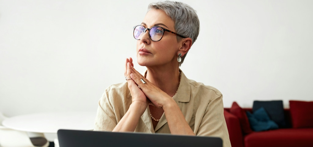 Older woman with short grey hair and glasses sitting thoughtfully at a laptop in her living room, hands clasped near her chin.