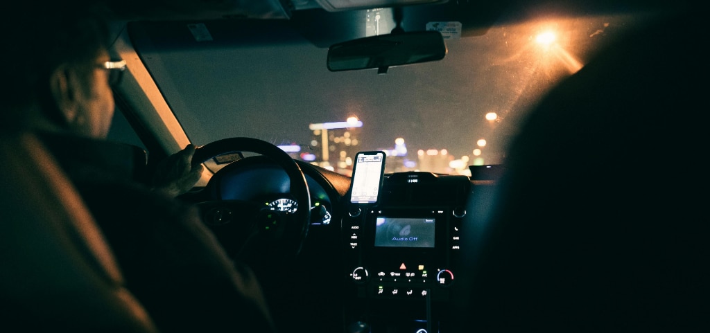 A back-seat view of a car interior showing the silhouettes of the driver and the passenger seat. There are glowing streetlights outside, and an illuminated phone and dashboard display inside.