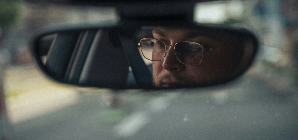 A reflection in the rear-view mirror shows a man driving in low light. He has light stubble and wears gold-rimmed rectangular glasses.