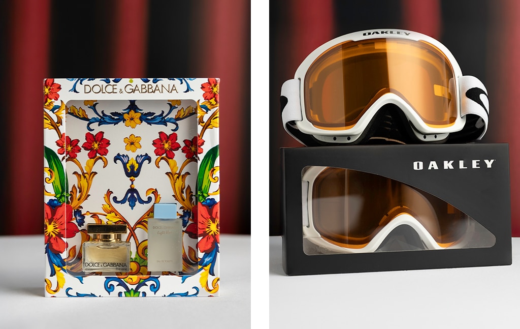 Two-image collage. First image: A Dolce & Gabbana duo perfume gift set on a red curtain backdrop. Second image: White and black Oakley ski goggles on top of a black Oakley box containing a matching pair of Oakley ski goggles, also on a red curtain backdrop.