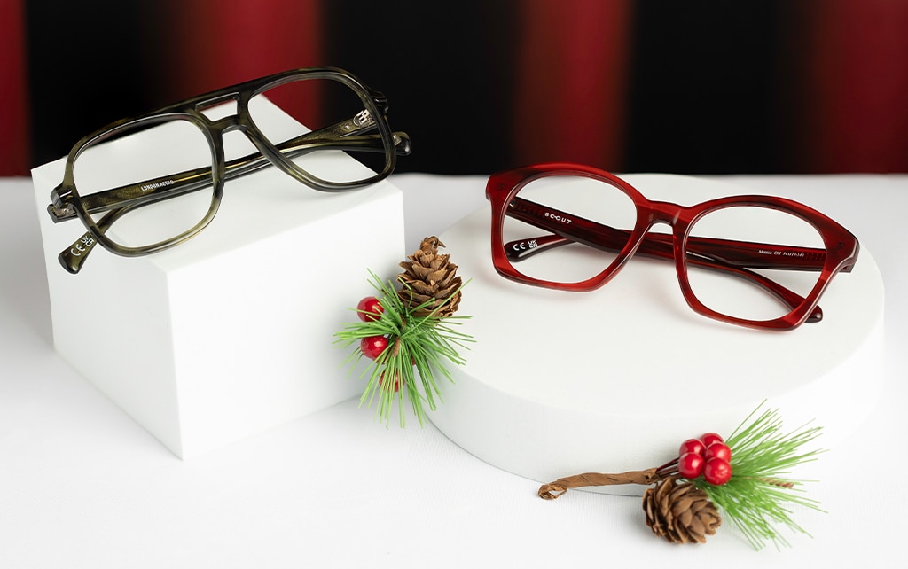 Two pairs of glasses displayed on a smooth white surface against a red curtain backdrop with green fern decorations featuring red berries and pine cones. One pair is a green acetate pilot frame resting on a white cube, and the other is a red square frame placed on a low white cylinder.