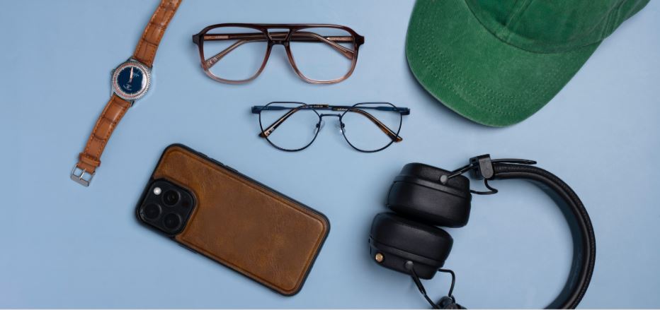 Two pairs of folded glasses, one metal and one acetate, resting on a light-blue background surrounded by a green baseball cap, black headphones, a mobile phone in a brown case, and a brown-strap watch.