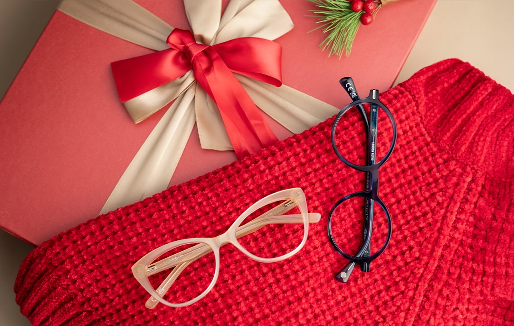 Two pairs of glasses—one round blue acetate frame and one pearly-coloured cat-eye frame—rest on a crimson-red jumper on top a red festive gift box topped with a gold-and-red bow and a small decorative fern with red berries.