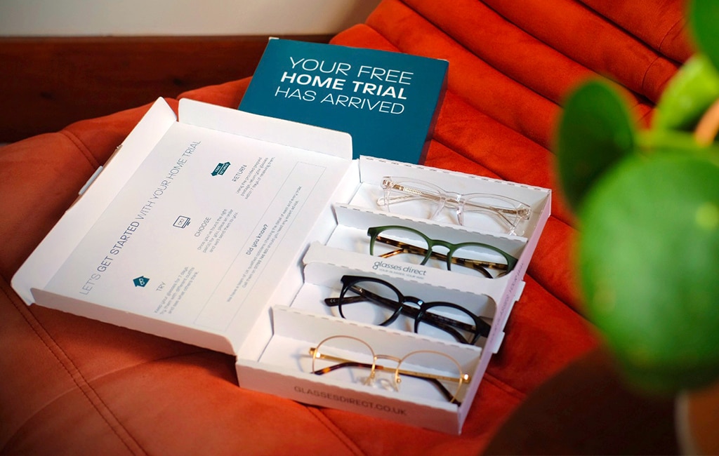An open Glasses Direct Home Trial pack displaying four different frame styles, resting on a red chair, with a green plant partially obscuring the right side of the scene.