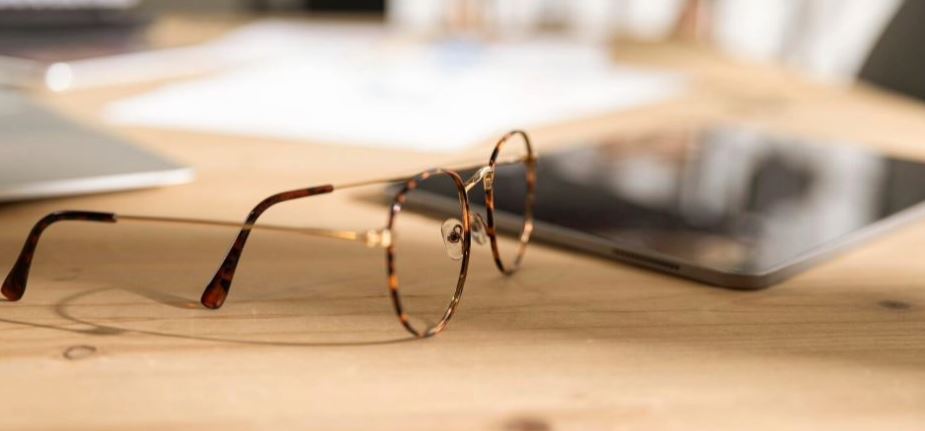 A pair of gold round glasses with tortoiseshell trim resting on a wooden table, with a black tablet and various items visible in the background. The lenses are clear of fog.