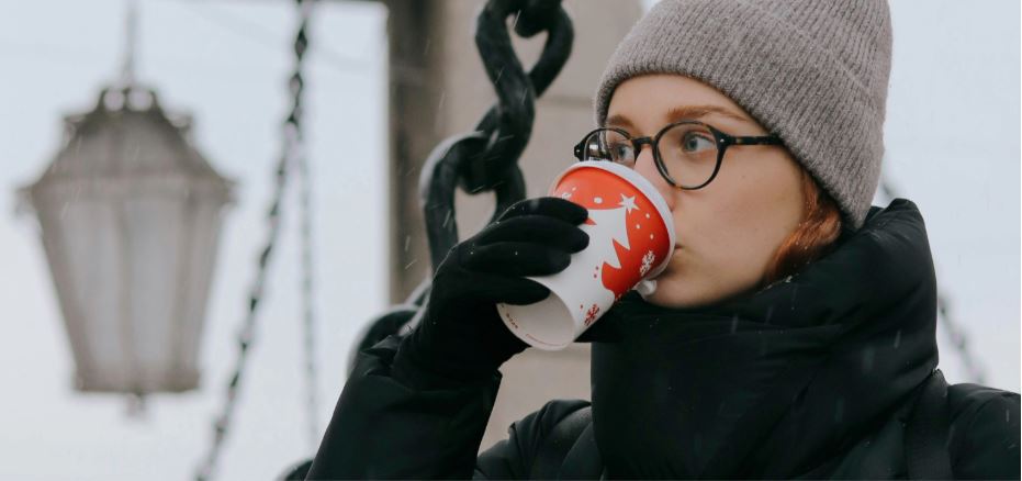 Close-up of a young woman wearing tortoiseshell glasses and a beanie, sipping from a Christmas-themed takeaway cup while dressed in a black winter coat and gloves, showing how to stop glasses from fogging when drinking hot drinks.