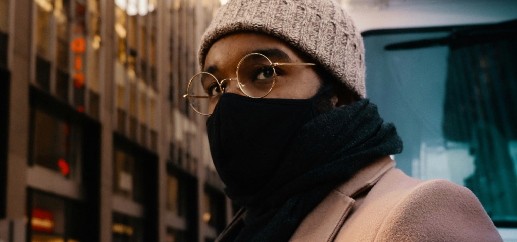 Close-up of a young man wearing gold-rimmed round glasses, a grey wool beanie, and a beige winter coat with a black scarf over his mouth, showing how to stop glasses from fogging up on a cold day in the city.