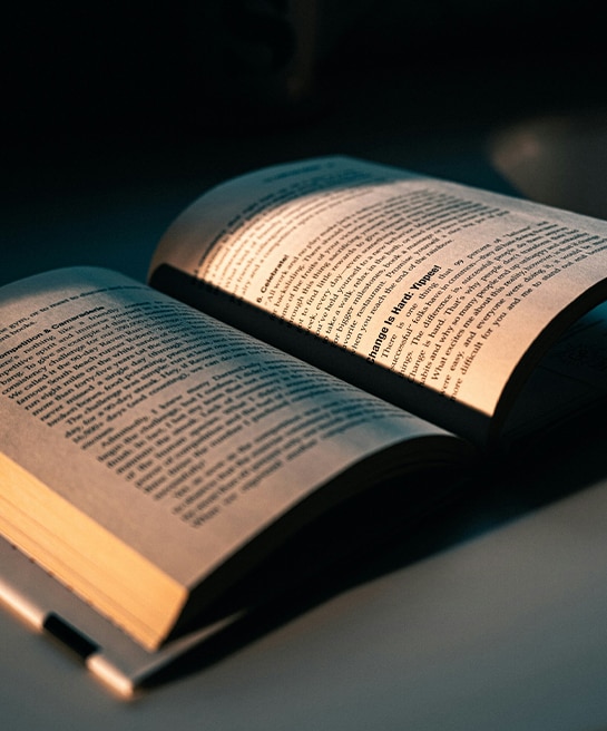 An open book resting on a flat surface in a dimly lit room, its pages softly illuminated against the darkness.