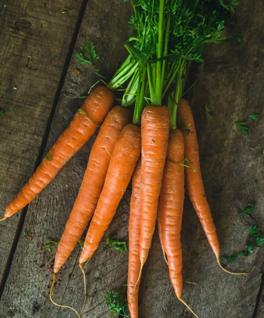 A bunch of fresh carrots with green tops resting on a wooden surface.