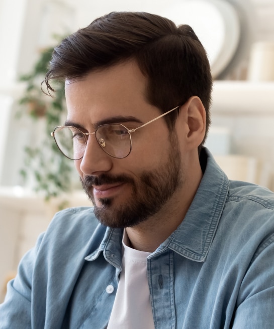 A close-up of a man with dark hair and a tidy beard, wearing gold-rimmed round metal glasses and a denim shirt over a white t-shirt. He smiles lightly in a bright, modern indoor setting.