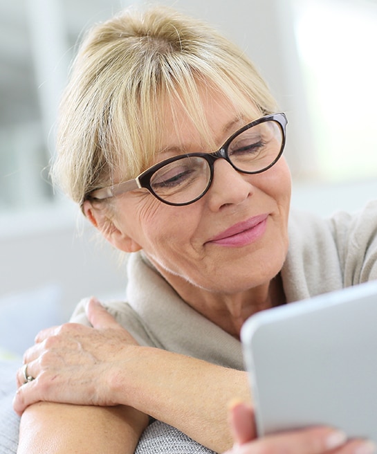 A close-up of an older woman with blonde hair, tied back, and a fringe. She is wearing black cat-eye glasses and a grey short-sleeve sweater, smiling as she holds a tablet in her right hand while her left hand rests on her right shoulder. She is in a bright indoor setting.