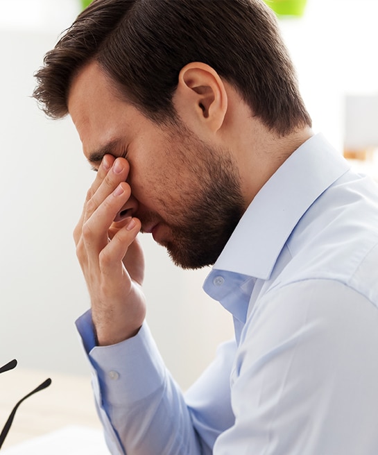A close-up of a young man with short dark hair and a beard, rubbing his eyes with his right hand in apparent discomfort while holding a pair of glasses in his left. He is in a bright, modern indoor setting.
