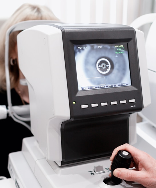 A close-up of an autorefractor machine, with a blonde woman partially visible behind it and a hand operating the device in the foreground.