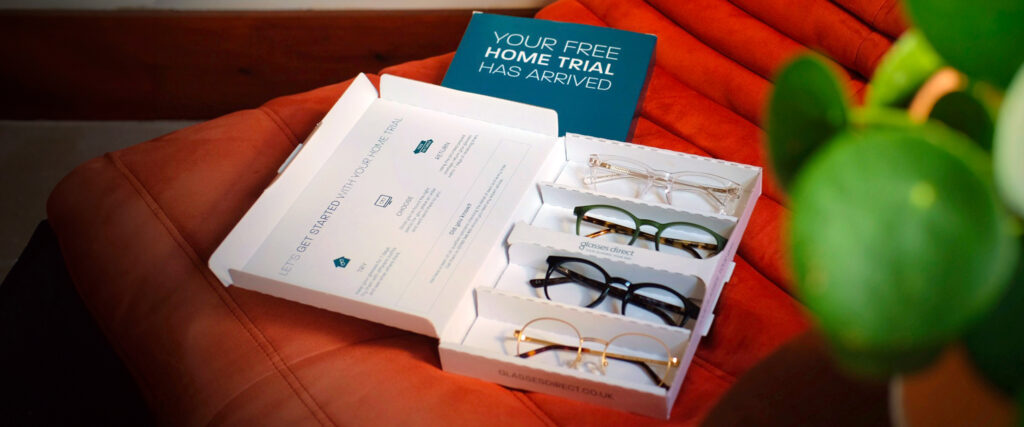 An open Glasses Direct Home Trial pack displaying four different frame styles, resting on a red chair, with a green plant partially obscuring the right side of the scene.