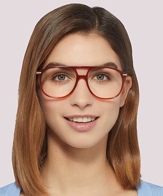 A woman facing the camera wears pilot-style orange acetate glasses and a light blue top. She has straight, shoulder-length brown hair and a soft, neutral smile against a pale background.