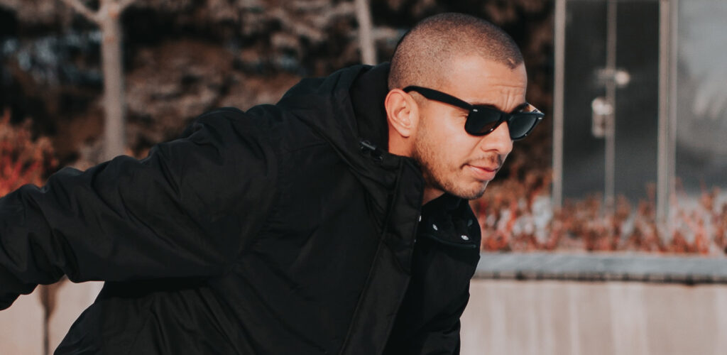 A close-up of a young man with a closely shaved head ice skating outdoors, leaning forward while wearing black Wayfarer-style sunglasses and a black padded jacket, with autumn foliage and an urban background softly blurred.