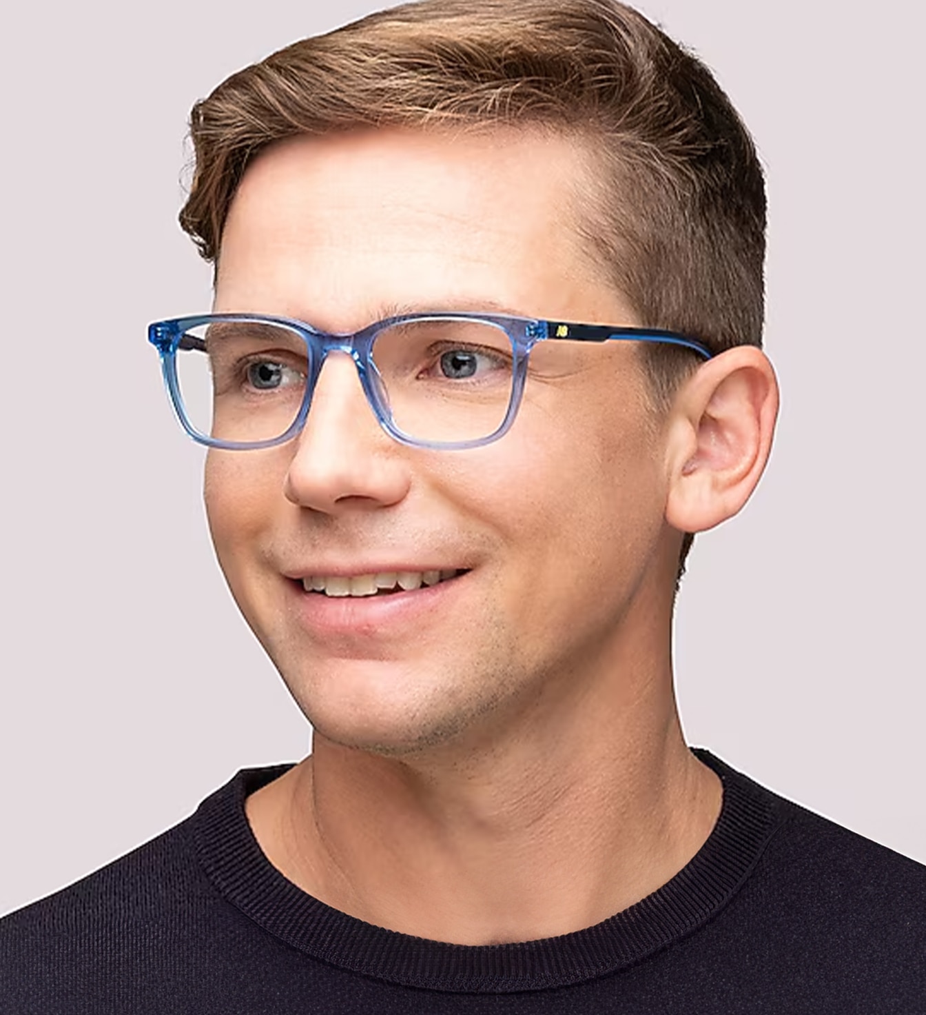 A close-up portrait of a man with a blonde quiff wearing transparent blue rectangular glasses and a black top against a plain background.
