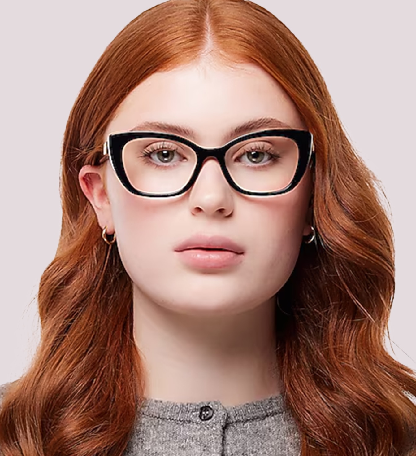 A close-up portrait of a woman, facing directly ahead, with long wavy red hair wearing bold black designer cat-eye glasses and a grey cardigan against a plain background.