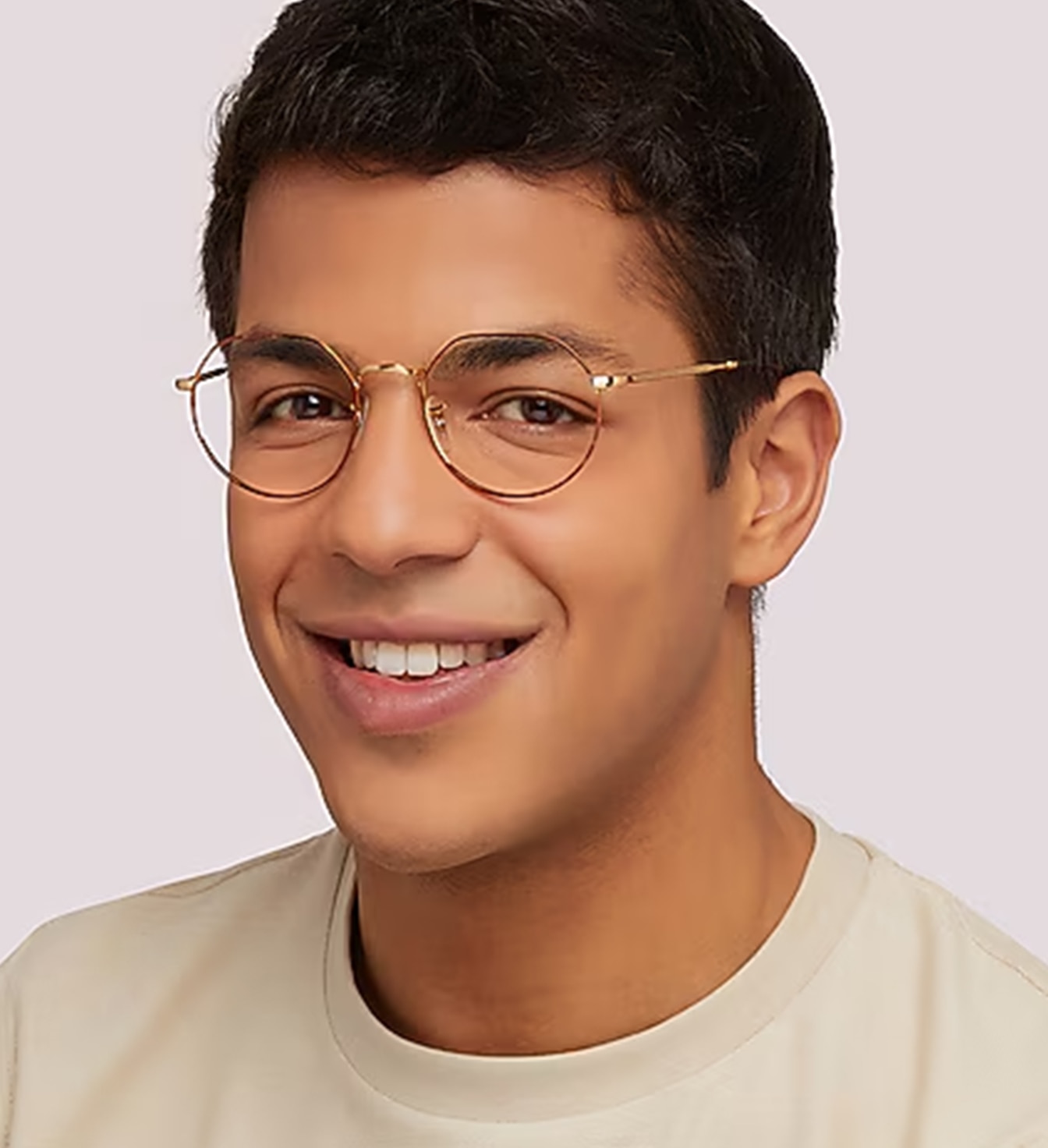 Close-up portrait of a smiling young man, positioned to his right, with short dark brown hair wearing circular gold Havana metal glasses and a cream t-shirt against a plain background.