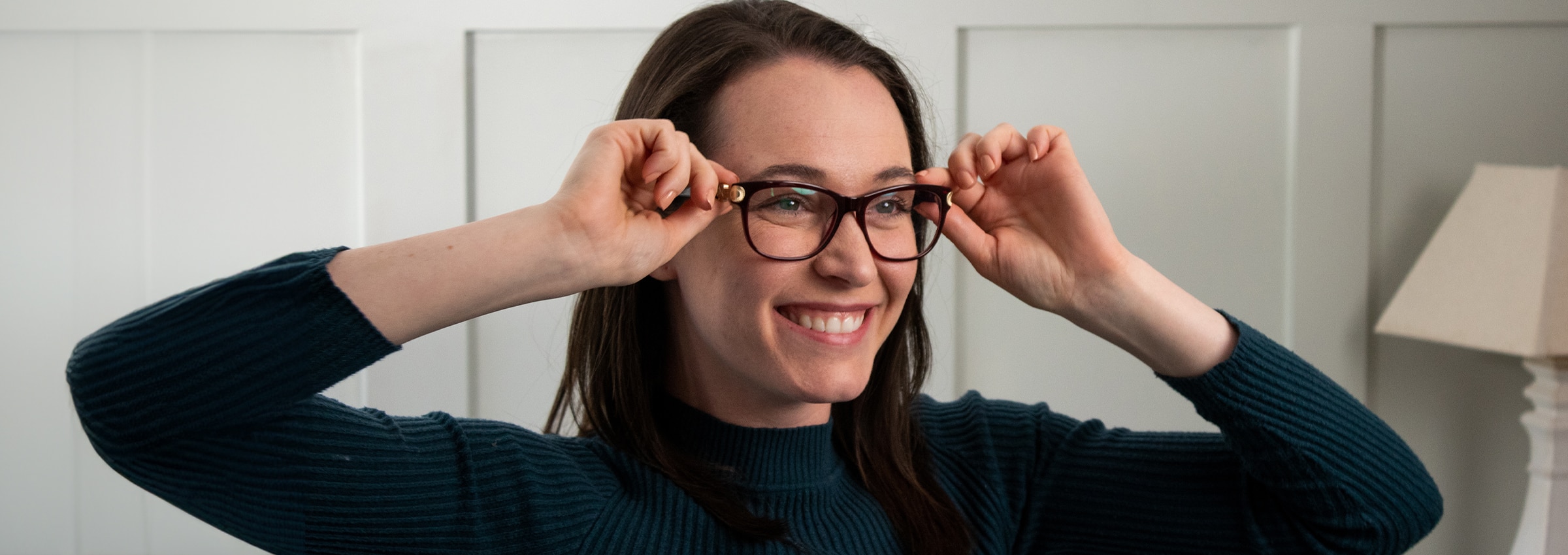 A woman smiling while adjusting her glasses at home, trying on frames in front of a mirror.