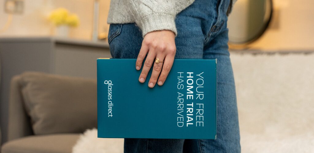 A person wearing a jumper and jeans holding a Home Trial box from Glasses Direct. A modern living room setting is blurred in the background. 
