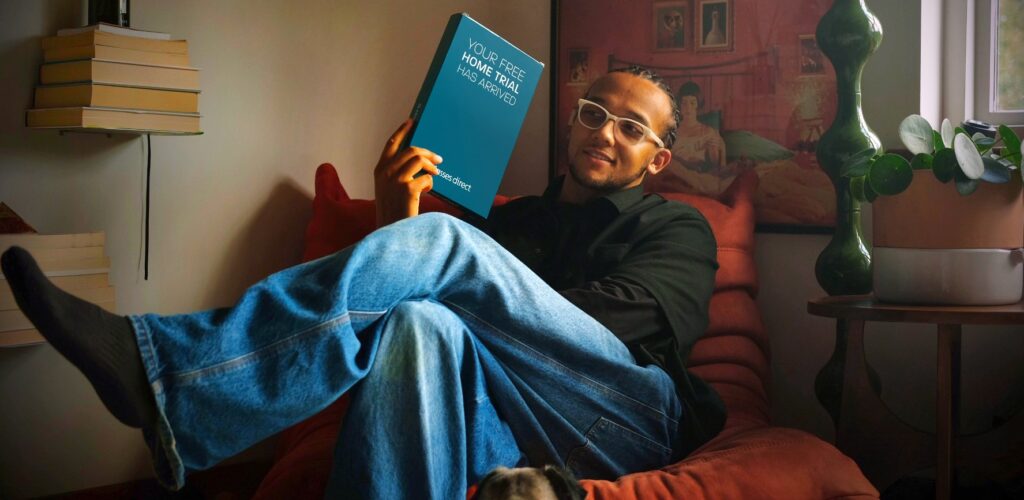 Perri Kiely sitting on a red chair in a living room setting, wearing a black shirt, blue jeans, and white eyeglasses, holding a Home Trial box.