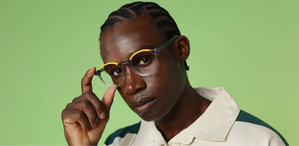 Young man wearing clear round glasses with a yellow brow detail, adjusting the frame against a green background, styled in a green and white polo shirt.