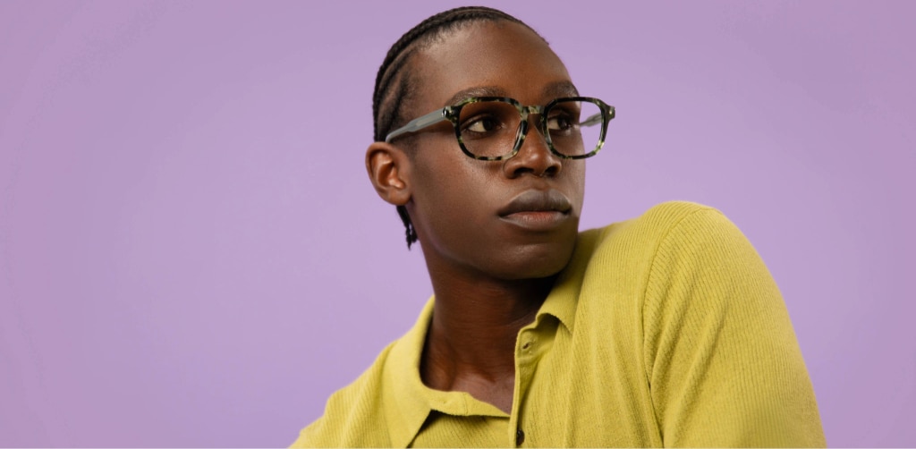 Man wearing green tortoiseshell rectangular glasses, looking to the side against a soft purple background, styled in a textured green shirt.