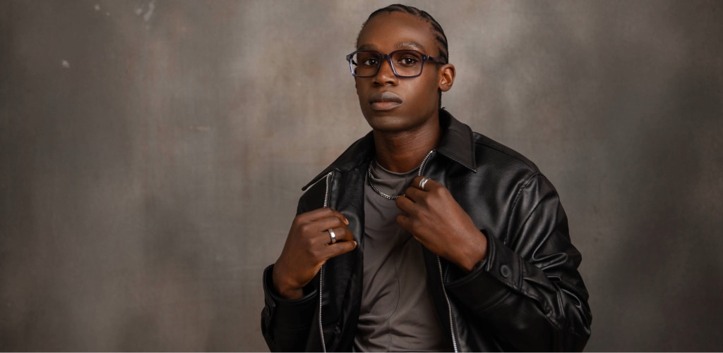 A young man wearing dark rectangular glasses and a black leather jacket over a grey t-shirt and silver-chain necklace, adjusting his collar while facing forward against a neutral studio background.