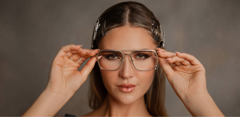 A woman wearing oversized metal-frame glasses looks directly at the camera while adjusting the frames with both hands. Her hair is styled with decorative clips, and she wears subtle makeup against a neutral background, highlighting a modern retro-inspired eyewear style.
