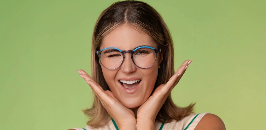 A woman wearing transparent blue-rimmed glasses smiles and winks at the camera, with her hands framing her face against a soft green background.