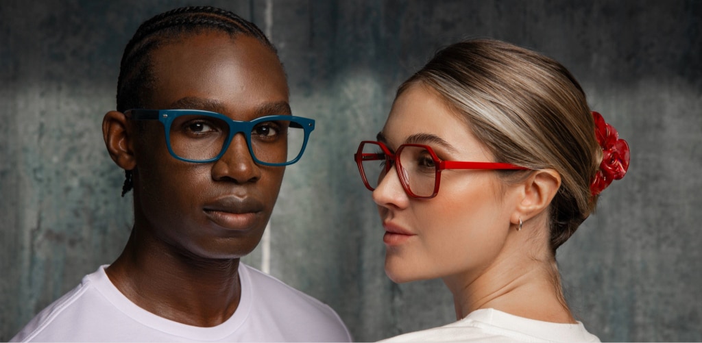 Two models wearing bold glasses pose against a grey neutral backdrop. The man wears oversized blue rectangular frames with a white t-shirt, while the woman wears red geometric frames with a white t-shirt, facing away from the camera with her head turned to the side.