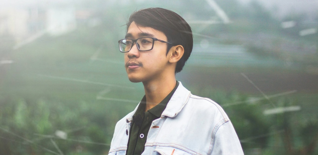 A man with short dark hair wearing black rectangular eyeglasses and a denim jacket over a dark shirt, standing outdoors in rainy or misty conditions. Despite the moisture in the air, the lenses appear clear, allowing his face to be seen sharply. The background is softly blurred with streaks of rain, highlighting improved visibility through the glasses compared to wet conditions.