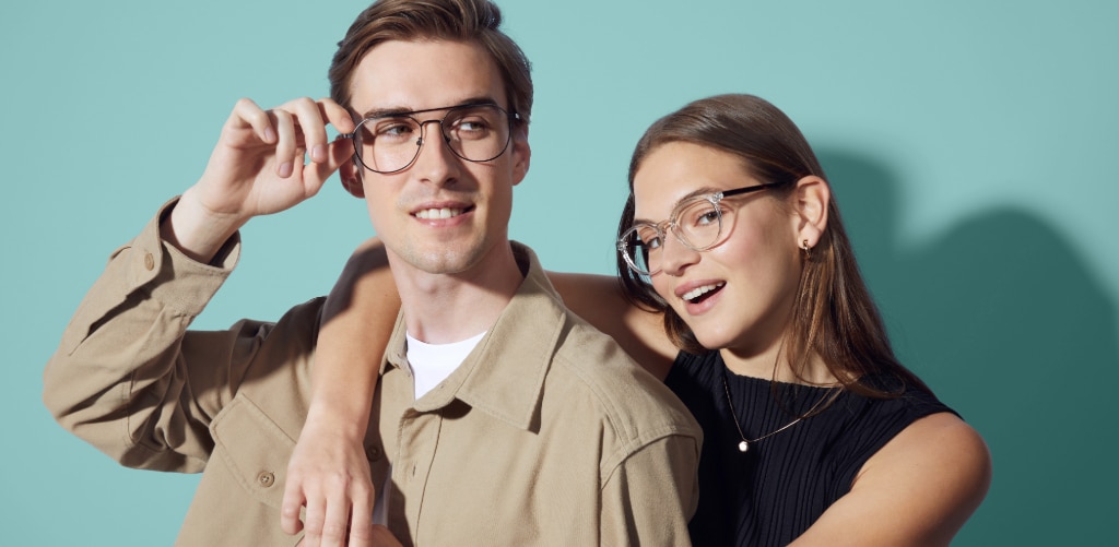 A young man with a blonde quiff, dressed in a brown jacket over a white tee and metal aviator glasses, stands beside a young woman with long, straight brown hair, wearing a black vest top and clear acetate frames. Set against a teal backdrop, he casually adjusts his glasses while she smiles warmly, her arm draped around his shoulder.