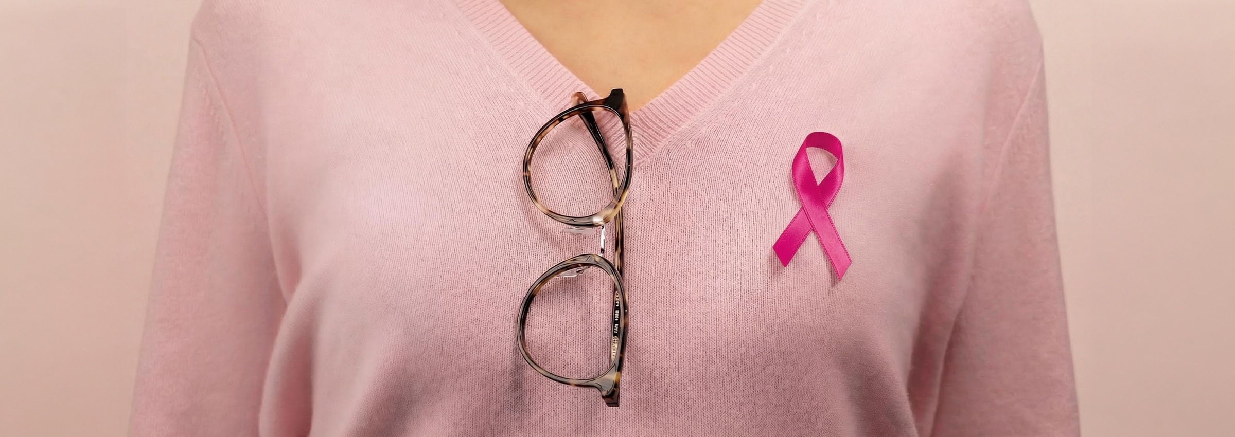 Close-up of a person wearing a soft pink sweater with tortoiseshell glasses hanging from the neckline and a pink breast cancer awareness ribbon pinned to the chest.