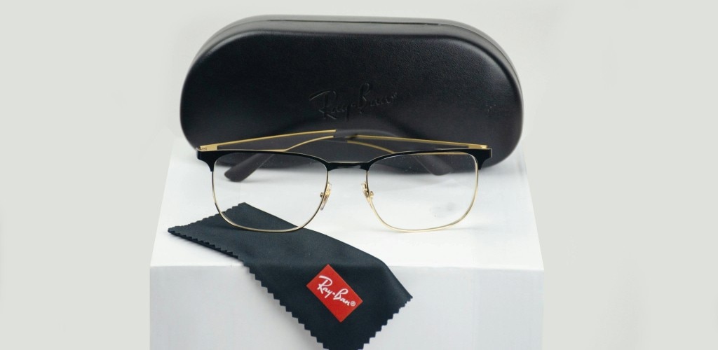 A pair of rectangular Ray-Ban eyeglasses with thin black-and-gold metal frames displayed on a white cube. Behind them is a black protective case, and in front lies a cleaning cloth with a visible Ray-Ban logo. The setup appears like a product display, highlighting the glasses along with their accessories.