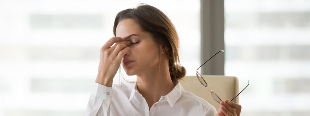 A young woman with brown hair tied loosely in a bun, holding a pair of glasses in one hand and rubbing her eyes with the other. She is wearing a white shirt, sitting on a cream chair in a bright indoor setting.
