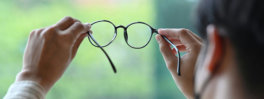 The back of a man with short dark hair holding a pair of round dark acetate frames up to a window and inspecting the lenses.