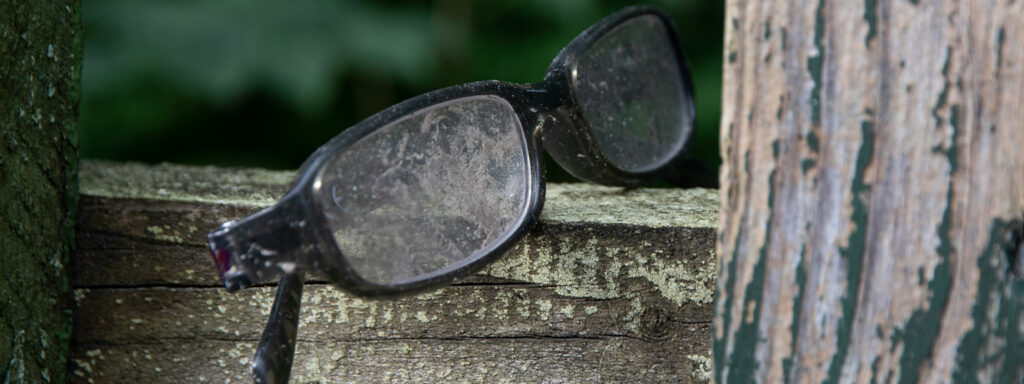 A pair of black rectangular acetate glasses covered in mud and dirt, folded up on a wooden fence.