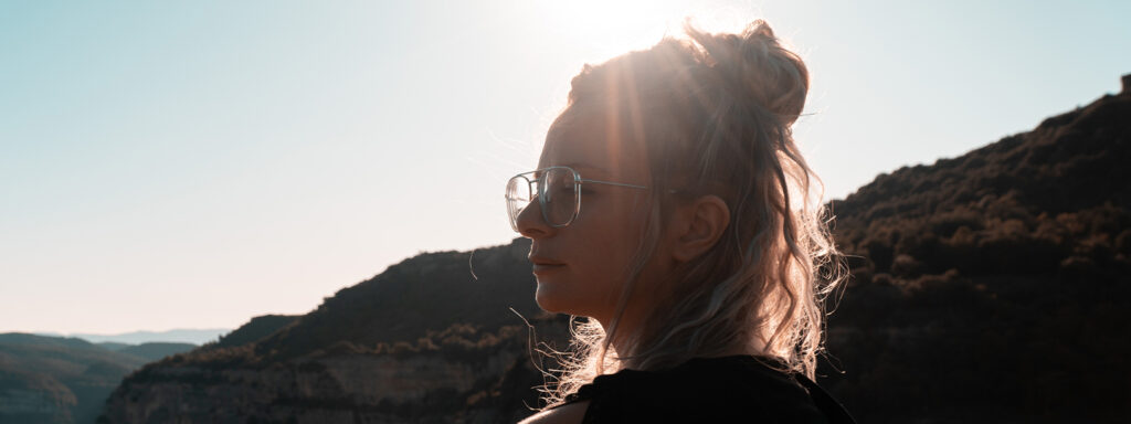A woman with part of her hair tied up in a bun, standing in the glare of sunlight, wearing metal aviator glasses and a dark jumper.