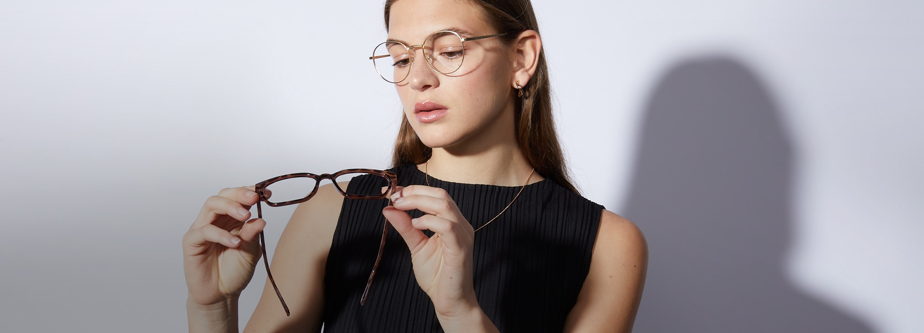 Woman with long straight brown hair wearing round gold metal glasses and a black sleeveless top while holding and inspecting another pair of tortoiseshell glasses.