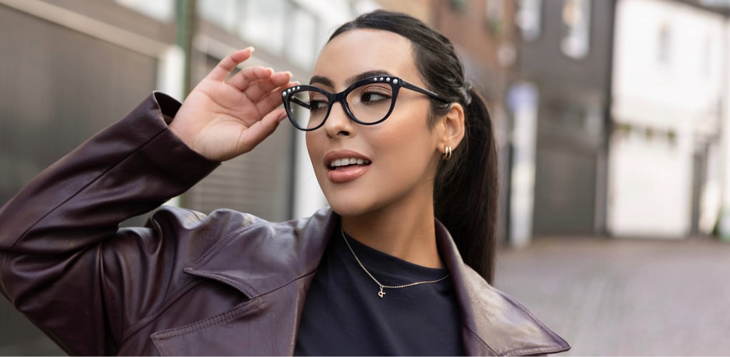 A woman wearing retro-inspired cat-eye glasses looks to the side while adjusting the frame. She is outdoors in an urban setting, dressed in a dark top and leather jacket, with her hair tied back and subtle gold jewellery visible.