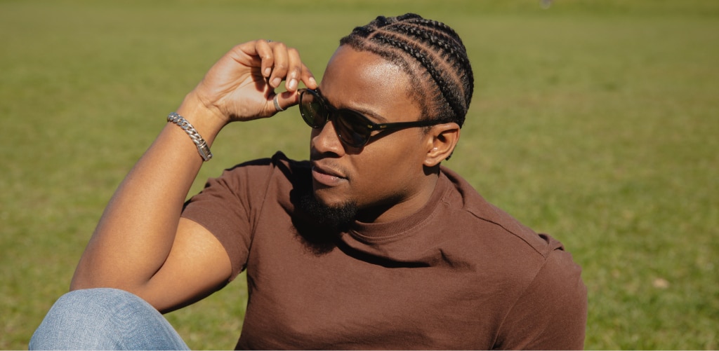 A young man with dark braided hair sits on grass in bright sunlight, adjusting his black sunglasses while wearing a brown t-shirt, blue jeans and a silver bracelet.