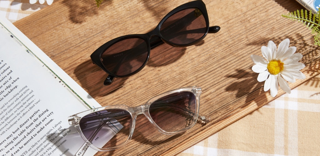 Two pairs of butterfly and cat-eye sunglasses—one black and one clear-framed—rest on a wooden board beside an open book and a white daisy, arranged in bright natural spring sunlight on a yellow checked blanket.