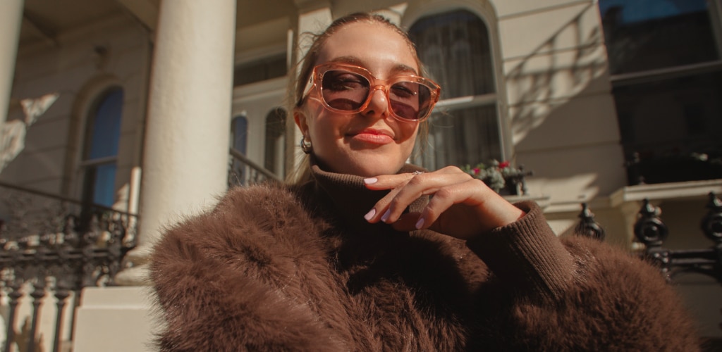 A young woman with her hair tied back, wearing clear pink sunglasses and a brown fluffy coat poses outdoors in bright sunlight, resting her hand under her chin, with white columns and a black iron fence in the background.