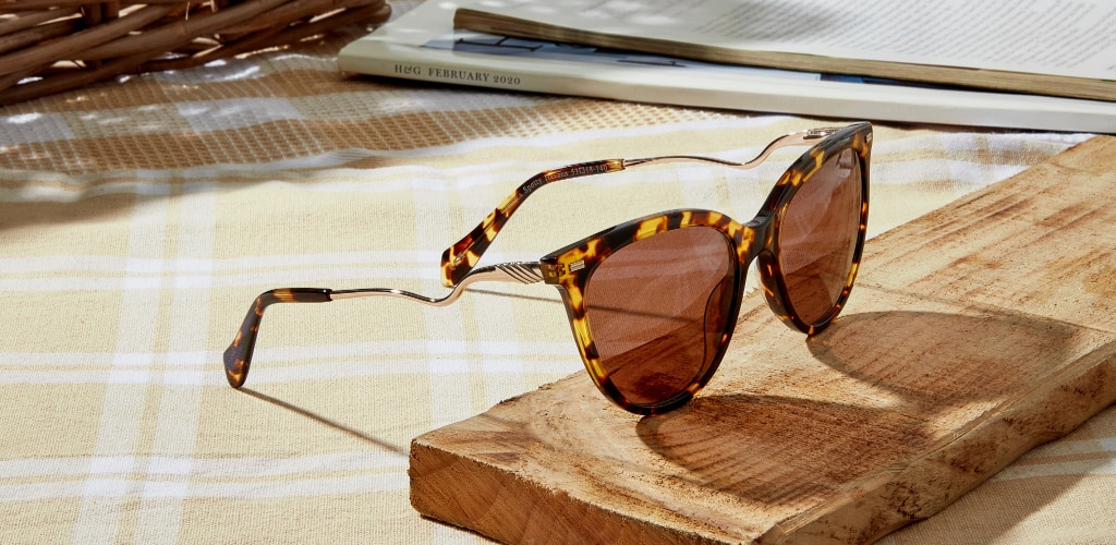 Tortoiseshell cat-eye sunglasses with gold curved metal temples and brown lenses rest on a small wooden block in bright sunlight, set on a yellow checked picnic blanket with magazines and a wicker basket in the background.