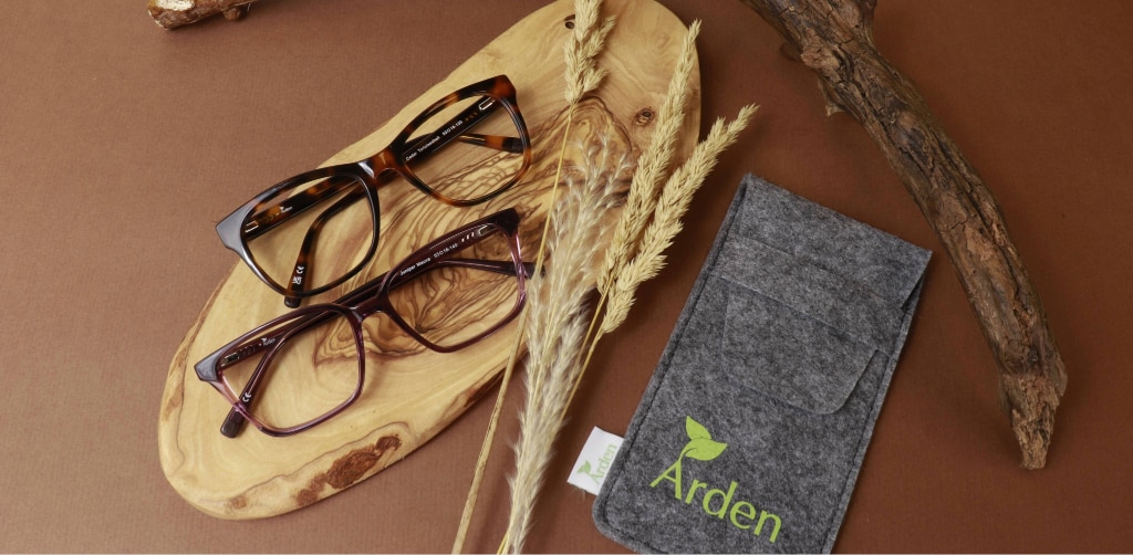 Two pairs of tortoiseshell glasses displayed on a wooden board on a brown background with natural elements, alongside an Arden-branded case, highlighting sustainable eyewear materials.