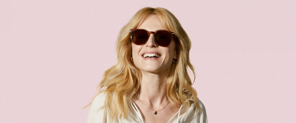 Smiling woman with long blonde wavy hair wearing brown tortoiseshell sunglasses against a soft pink background, styled for a Mother’s Day gift guide.